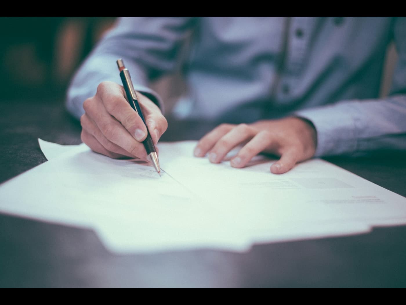 Unas manos escriben con pluma sobre un documento impreso, apoyado en una mesa de trabajo